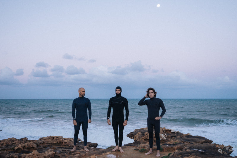 Surfer in a wetsuit seen from behind walking up from the water with a foam board