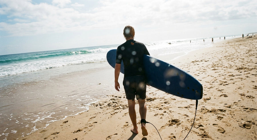 Surfeur de dos marchant vers l’eau avec sa planche en mousse sous le bras